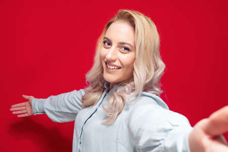 Selfie. Close up pleased beautiful girl takes photo of himself with her smartphone, isolated on red background. Focus on face. Studio shotの写真素材