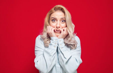 Scared girl biting fingernail,being terrible accident, isolated on red background. Frightened woman staring to you. Studio shotの写真素材