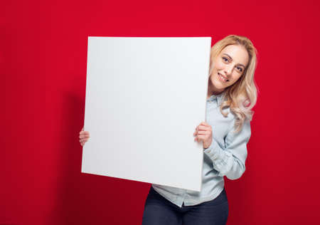 Smiling young women holding white board, isolated on red background. One cute girl showing blank empty paper billboard with blank space for text. Studio shotの写真素材