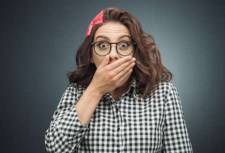 Astonished woman covers her mouth over dark gray - black background. Surprised girl. Unbelievable concept. Studio shotの写真素材