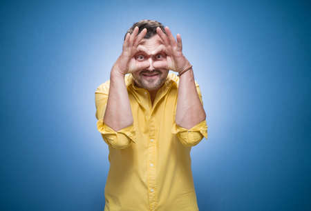 Silly guy staring to you over blue background, dresses in yellow shirt. Half length funny young man. Joke concept. Studio shotの写真素材