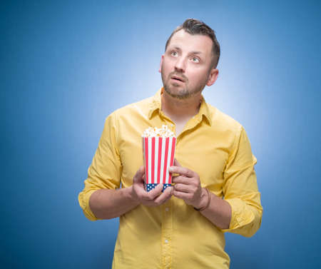 Shocked guy with popcorn over blue background, dresses in yellow shirt. Frightened young man with astonished expression on his face. Weekend times - movies watching concept. Studio shotの写真素材