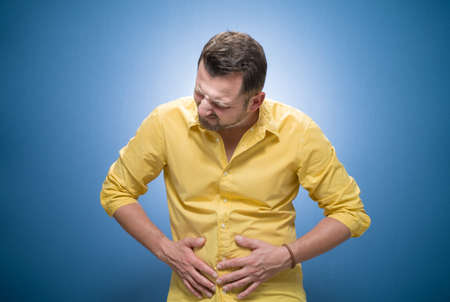 Young man suffering from stomachache - stomach ache over blue background, dresses in yellow shirt. Ill guy with chronic gastritis. Studio shotの写真素材