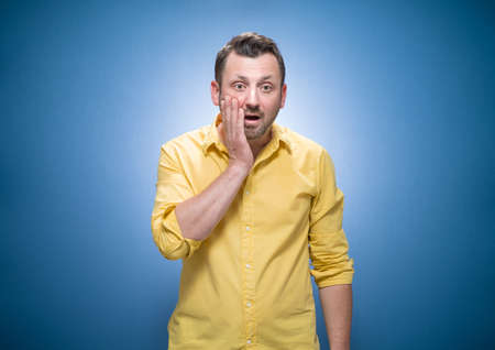 Oops. Ashamed young man with hand on cheek. Shame concept over blue background, dresses in yellow shirt. Studio shotの写真素材