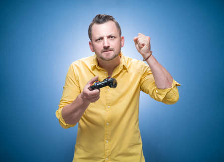 Angry man feeling upset after loosing video game, frustrated and aggressive guy shows fist, using joystick over blue background, dresses in yellow shirt. Studio shotの写真素材