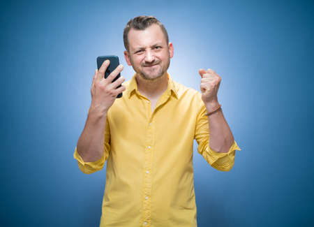 Nervous man showing her mobile phone over blue background, dresses in yellow shirt. Furious guy shows smartphone. Studio shotの写真素材