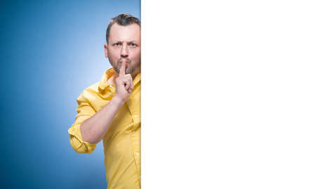 Hush. Man holding finger on lips, keep the secret concept over blue background, dresses in yellow shirt. Studio shotの写真素材