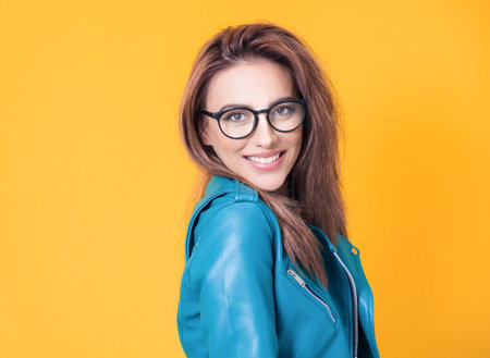 Face of nice girl keeps chin on shoulder, wearing blue leather jacket, isolated on yellow background. Closeup portrait of a cheerful woman . Studio shotの写真素材