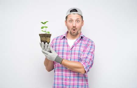 Astonished male gardener holding plant in soil with plant in her hands. Plant care concept, isolated on white background. Amazed male farmer. Studio shotの写真素材