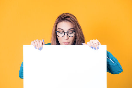 Close-up woman behind white board and looking down to empty space, wearing blue leather jacket, isolated on yellow background. Pretty girl showing blank space for text near her head. Studio shotの写真素材