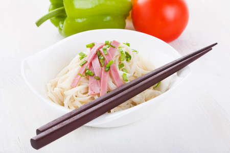 Tasty noodles with ham green onions in a beautiful white bowl on a white wooden background. Peppers and tomatoes.の写真素材