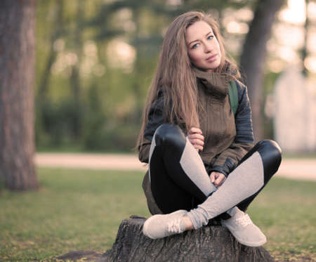 The Beautiful Young Girl with Long Hair, in a Gacket sits on a Stump in the Summerの写真素材