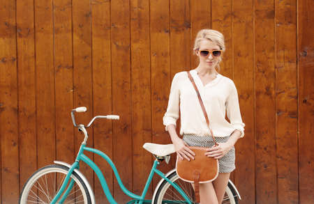 Young sexy blonde girl is standing near the vintage green bicycle with brown vintage bag in orange sunglasses.の写真素材