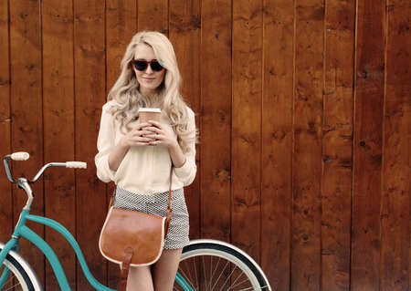 Young sexy blonde girl with long hair with brown vintage bag in sunglasses standing near vintage green bicycle and holding a cup of coffee, have fun and good mood looking in camera and smilingの写真素材