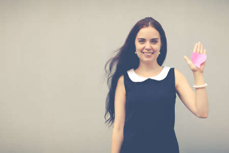 Beautiful brunette with long hair near the wall on the street in the summer. On a raised hand red paper heartの写真素材