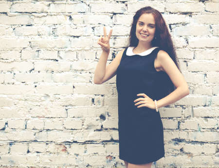Beautiful brunette with long hair in a short black showing two fingers up near on background of white brick wallの写真素材