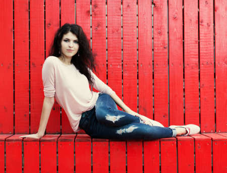 Beautiful tall girl with long hair brunette in jeans sits near wall of red wooden planksの写真素材