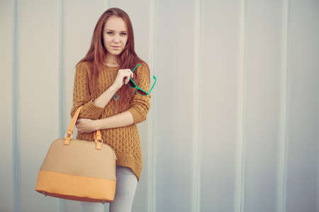 Beautiful redheaded girl with fashionable big bag standing near gray wallの写真素材
