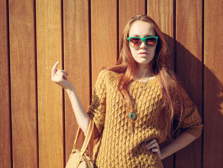 Beautiful redheaded girl with fashionable big bag in sunglasses standing near wooden wallの写真素材