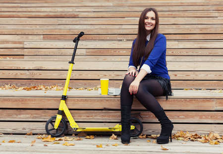 Beautiful brunette sitting on wooden steps with a glass and donuts. Yellow scooter stands nextの写真素材