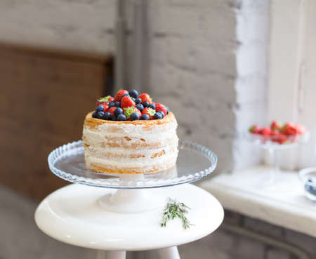 Beautiful biscuit cake with white cream decorated with strawberries and blueberries on a white pedestal stands at the windowの写真素材