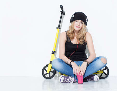 Young beautiful cheerful fashion girl in jeans, sneakers, hat sits on a yellow scooter and listening to music on headphones. Keeps cool pink glass in handの写真素材