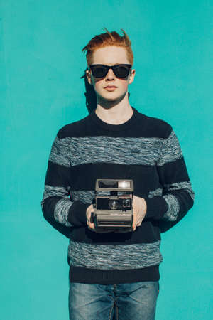 Young redhead man in a sweater and jeans and sunglasses standing next to turquoise wall and taking photos vintage camera warm summer sunny dayの写真素材