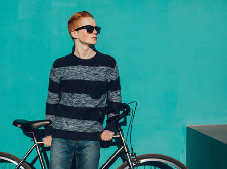 young redhead man standing next to a vintage bicycle in suglasses near turquoise wall warm summer sunny dayの写真素材