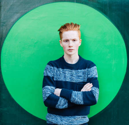 Young redhead man in a sweater and jeans standing next to green wall with folded arms