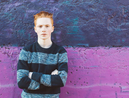Young redhead man in a sweater and jeans standing next to purple wall with folded armsの写真素材