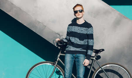 Young redhead man standing next to a vintage bicycle near turquoise wall warm summer sunny dayの写真素材