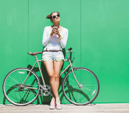 Pretty girl in shorts and t-shirt and sunglasses stands with  bicycle fix gear nex to green wall bright sunny dayの写真素材