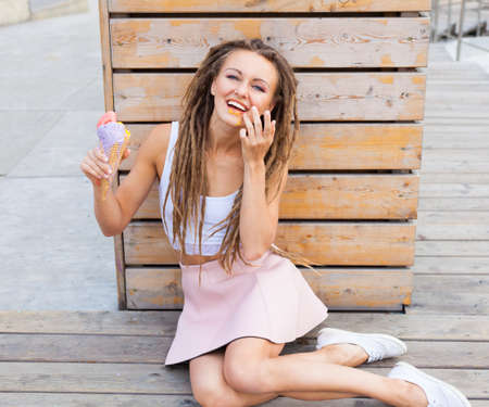 beautiful girl with dreadlocks in pink skirt sitting on the veranda and eating colorful ice-cream cone on a warm summer evening.の写真素材