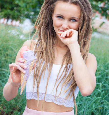 Young sexy blonde girl with dreads eating multicolored ice cream in waffle cones in summer evening, ...