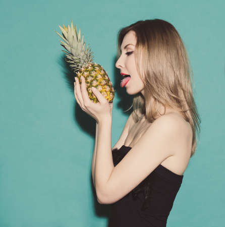 girl holding hands pineapple and reaches for his tongue in a black dress on a green background in the studioの写真素材