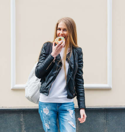 Beautiful girl bites a delicious donut walking on the streets of European city. Warm spring. Outdoor