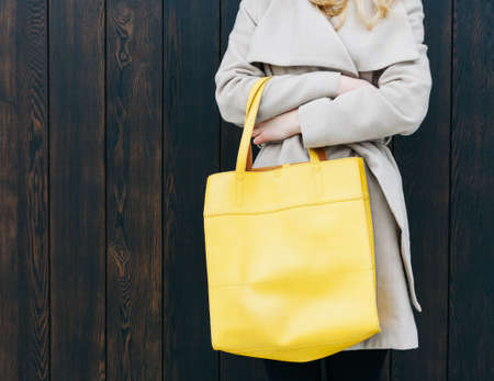 Fashionable beautiful big yellow handbag on the arm of the girl in a fashionable coat, posing near the wall. Part of body. Warm color.の写真素材