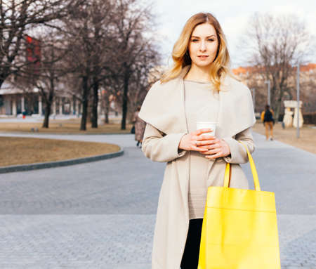 Young beautiful pretty girl walking along the street with big yellow handbag and cup of coffee. Europe city. Warm color. Outdoor.の写真素材
