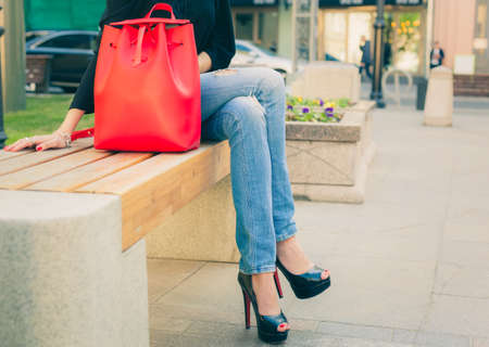 Girl in jeans and a fashionable high-heeled shoes sitting warm summer evening on the bench in the european city. Part of body. Warm color. Outdoor.の写真素材