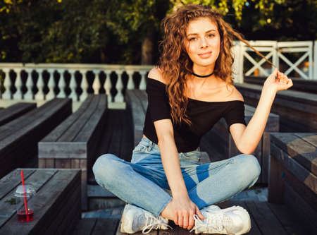 Beautiful long-haired girl sitting in a park on a summer evening. Dressed in a black top, sneakers and jeans. Beautiful spring sunshine.の写真素材