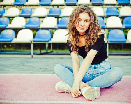 Outdoor Summer Fashionable sexy sensual portrait of a beautiful brunette girl with long hair posing in black top and jeans, having fun near the stadium. Youth and beauty. On the Sunset.の写真素材