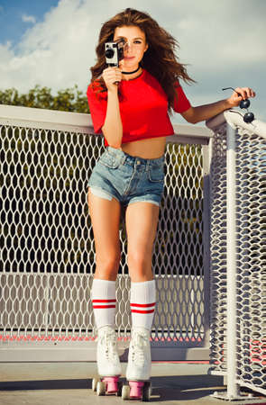 Young blonde happy smiling surprised girl in red top, shorts, sunglasses and white spats and roller derby skates on the stairs. Posing holding a vintage movie camera. Outdoor.の写真素材