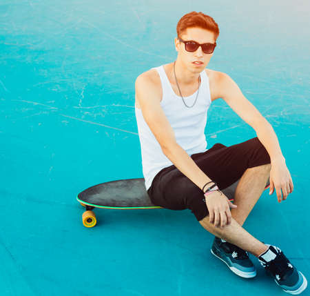 Fashionable young man sitting on a longboard in the ramp for skateboarding in the summer. A skateboarder wearing white shirt, shorts and sneakers. Outdoor.の写真素材