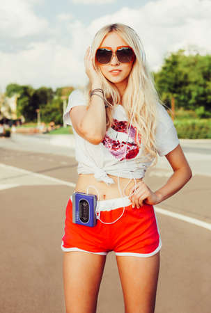 Summer fashion stylish portrait of young pretty sexy blonde girl posing in vinage sunglasses, T-shirt, shorts and listening to music with headphones on vintage cassette player. Outdoor.の写真素材