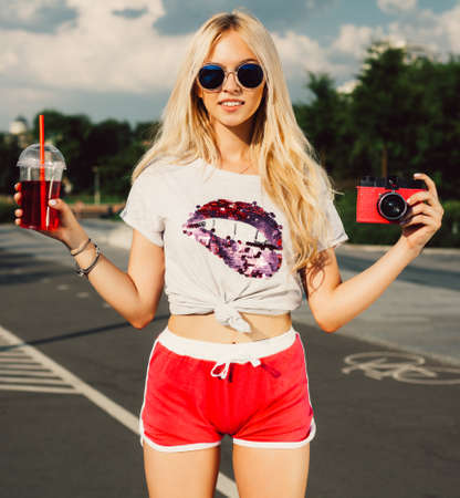 Beautiful young blond woman posing with a drink and a vintage red camera hot summer day. Outdoor.の写真素材