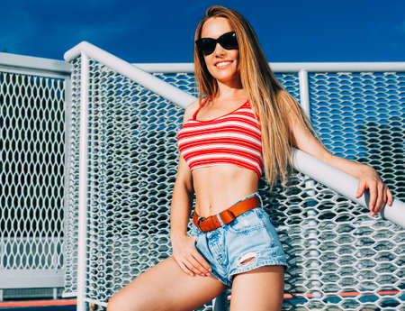 Beautiful young blond sexy woman standing on the stairs in shorts, t-shirt with sunglasses. Warm...