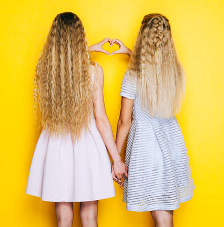 Love and friendship forever. Two girls girlfriends standing back and show making heart sign. Indoor. On yellow backgrownd.の写真素材