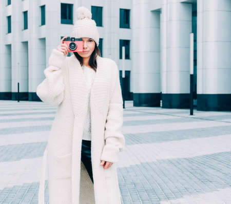 Beautiful fashionable girl in a knit coat on the street European city taking pictures pink vintage camera. Streetstyle.の写真素材
