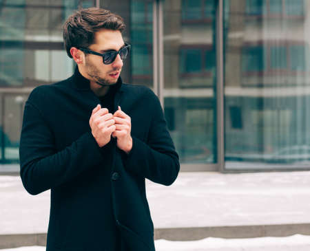 Portrait of young trendy man wearing a black coat with a raised collar in sunglasses. Street style. On the background a business building. Fashion. Close up.の写真素材
