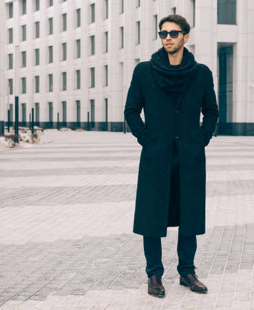 Fashion and Style. A young man in a fashionable black coat and sunglasses posing near the business center. Outdoor.の写真素材